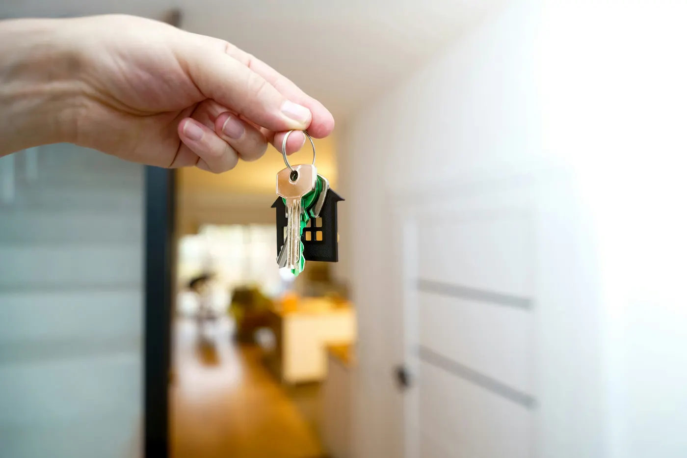 A hand holding up a set of house keys on a small house-shaped keyring, in the doorway of a bright, empty home
